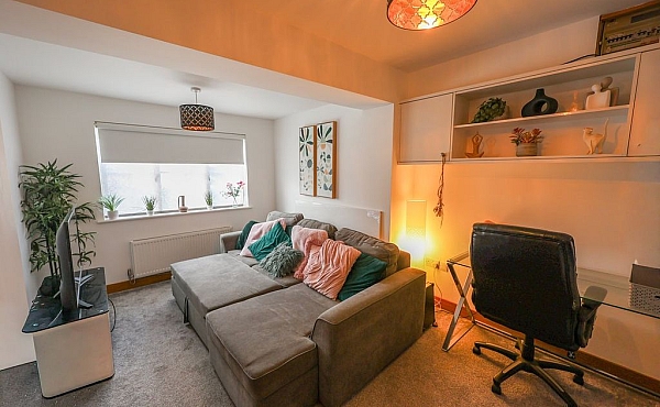 Room: bedroom | A well-lit indoor den featuring a sofa bed, loveseat, chair, potted plant, and decorative lampshade. | Accessibility: Photograph showing a well-lit indoor den with a neutral-colored sofa bed, a matching loveseat and armchair, a potted plant, and a decorative lampshade, all set on a wooden floor with white walls and ceiling lighting.