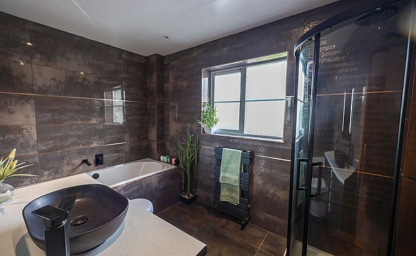 Room: Bathroom | A well-maintained bathroom featuring a sink, bathtub, mirror, potted plant, and a door, with tiled walls and a window for natural light. | Accessibility: Photograph showing a bathroom with tiled walls, a white porcelain sink with a chrome faucet, a built-in bathtub, a rectangular mirror above the sink, a small potted plant on the countertop, a wooden door, and a window providing natural light.