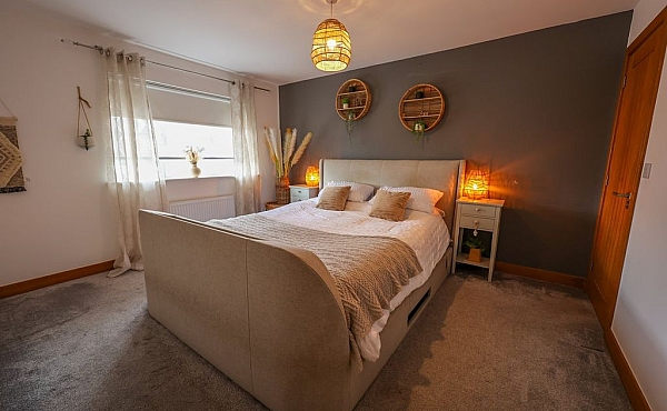 Room: bedroom | A well-presented bedroom featuring a bed with linens, pillows, a duvet cover, a nightstand, and a clean, inviting interior. | Accessibility: Photograph showing a bedroom with a neutral-colored bed dressed in white linens, pillows, and a duvet, a wooden nightstand beside the bed, and soft ambient lighting illuminating the room's light-colored walls and hardwood floor.