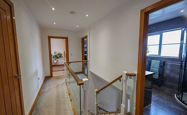Room: First Floor Hall | A well-maintained indoor room featuring a chair, with visible wall, door, ceiling, and flooring details. | Accessibility: Photograph showing a well-maintained indoor room with light-colored wooden flooring, a dark-colored upholstered chair, white painted walls, a wooden door, and ceiling-mounted lighting fixtures.