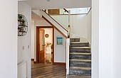 Room: Ground Floor Hall | An indoor staircase with a wooden handrail and balusters, featuring detailed molding and a clean, well-maintained floor. | Accessibility: Photograph showing an indoor staircase with a polished wooden handrail and matching balusters, detailed molding along the wall, and a clean, light-colored tiled floor illuminated by natural light.