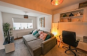 Room: bedroom | A well-lit indoor den featuring a sofa bed, loveseat, chair, potted plant, and decorative lampshade. | Accessibility: Photograph showing a well-lit indoor den with a neutral-colored sofa bed, a matching loveseat and armchair, a potted plant, and a decorative lampshade, all set on a wooden floor with white walls and ceiling lighting.