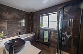 Room: Bathroom | A well-maintained bathroom featuring a sink, bathtub, mirror, potted plant, and a door, with tiled walls and a window for natural light. | Accessibility: Photograph showing a bathroom with tiled walls, a white porcelain sink with a chrome faucet, a built-in bathtub, a rectangular mirror above the sink, a small potted plant on the countertop, a wooden door, and a window providing natural light.