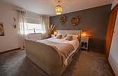 Room: bedroom | A well-presented bedroom featuring a bed with linens, pillows, a duvet cover, a nightstand, and a clean, inviting interior. | Accessibility: Photograph showing a bedroom with a neutral-colored bed dressed in white linens, pillows, and a duvet, a wooden nightstand beside the bed, and soft ambient lighting illuminating the room's light-colored walls and hardwood floor.