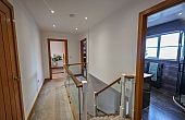 Room: First Floor Hall | A well-maintained indoor room featuring a chair, with visible wall, door, ceiling, and flooring details. | Accessibility: Photograph showing a well-maintained indoor room with light-colored wooden flooring, a dark-colored upholstered chair, white painted walls, a wooden door, and ceiling-mounted lighting fixtures.